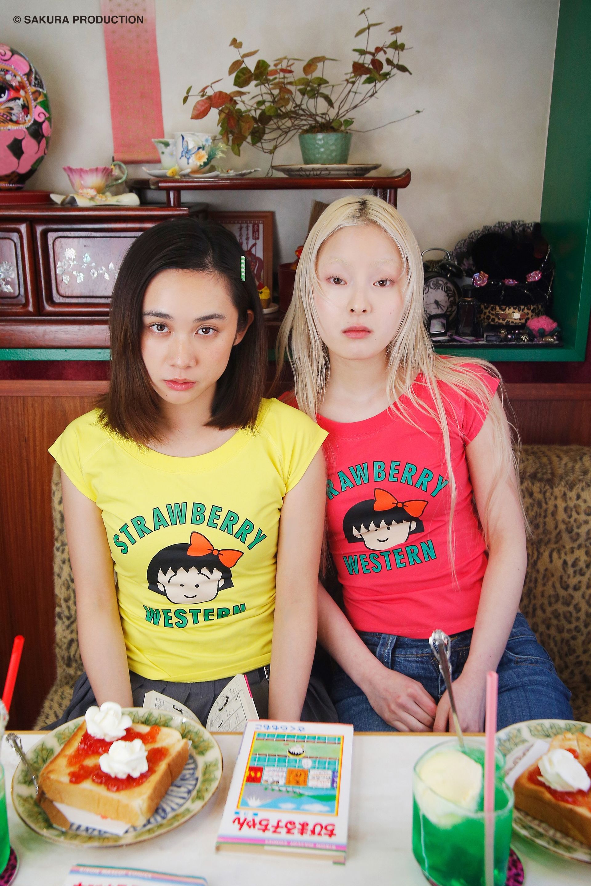 Two girls sitting in a Japanese cafe. The girl on the left wears the Yellow Chibi Maruko Chan Raglan Top and the girl on the right wears the Hot Pink version.