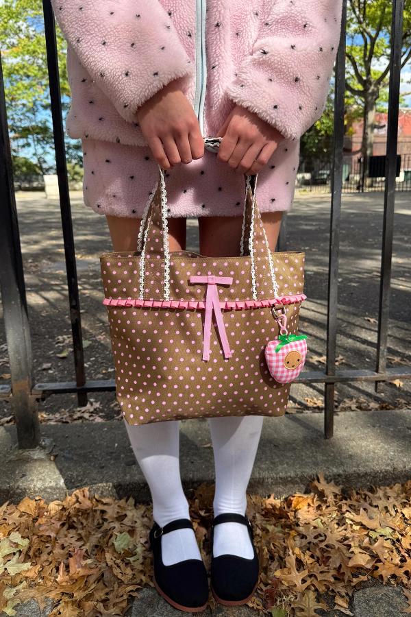Person standing outdoors holds a small brown shoulder tote bag with white polka dots, trimmed with a pink ribbon and lace detail.