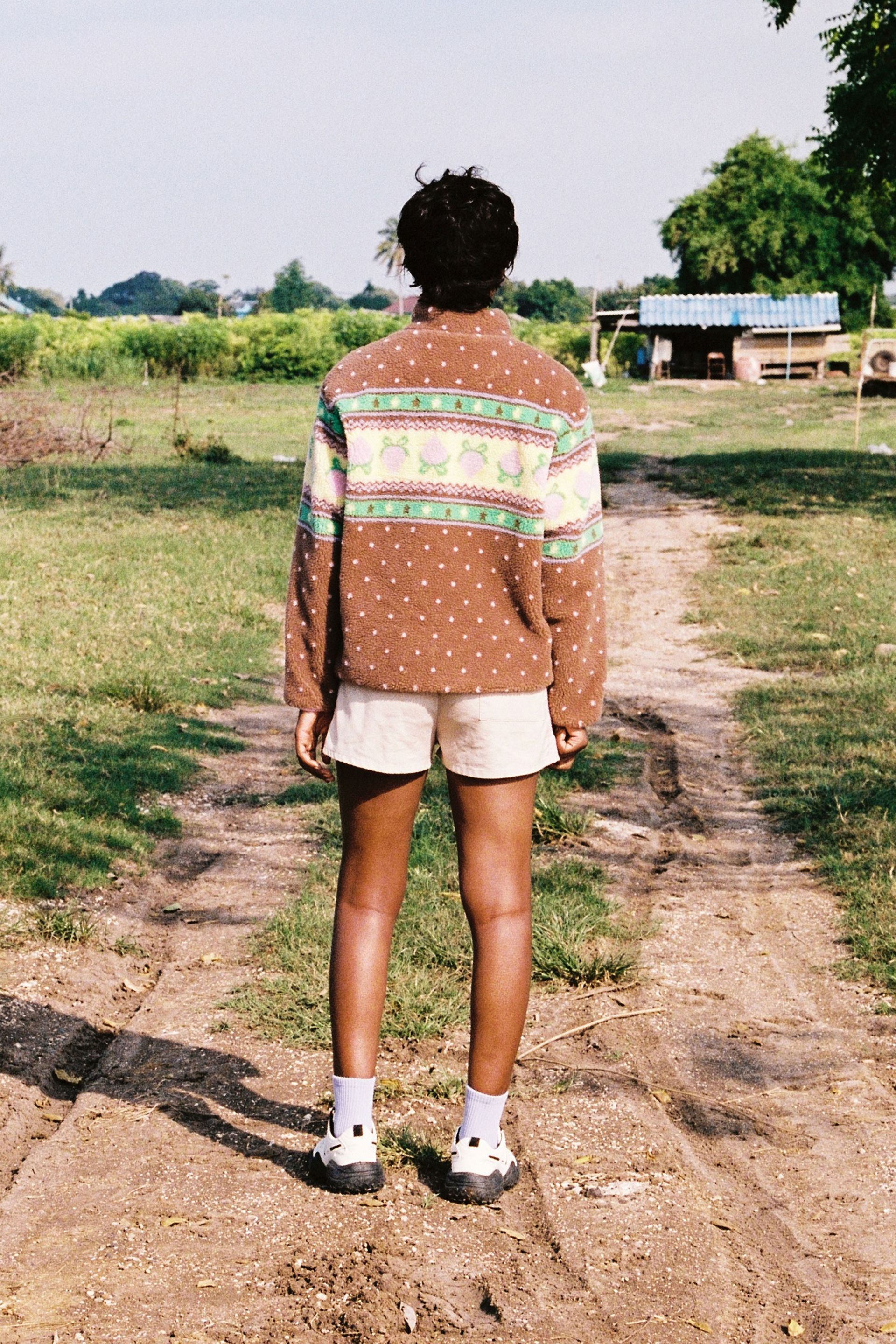Back of a person at a farm in the Brown fluffy jacquard knit sherpa jacket in a choco strawberry border motif with binding, zippers and a metal heart shape zipper pull.