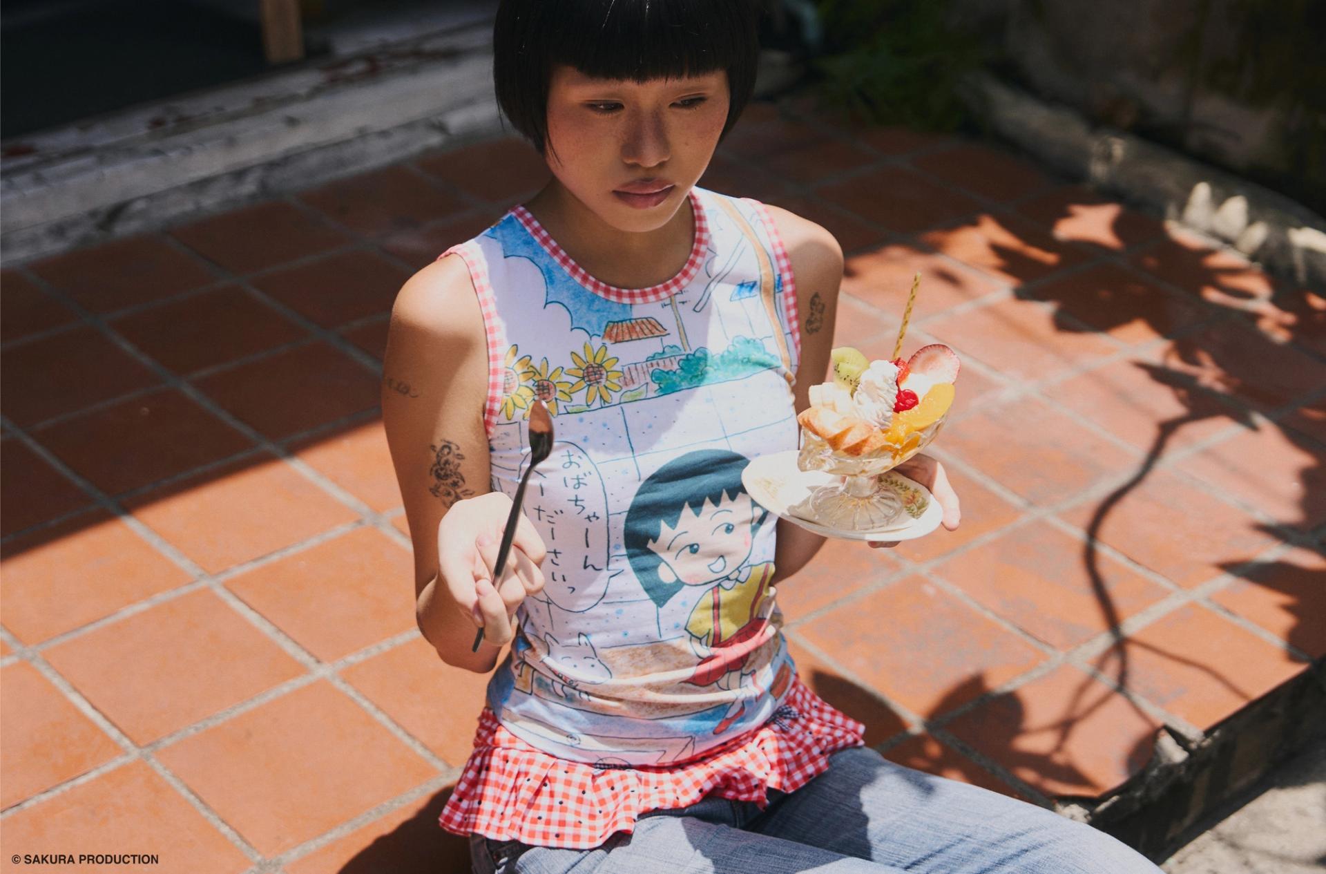Woman sitting on stoop with a parfait wearing a tank top with a print of the original Chibi Maruko Chan manga cover art illustration.