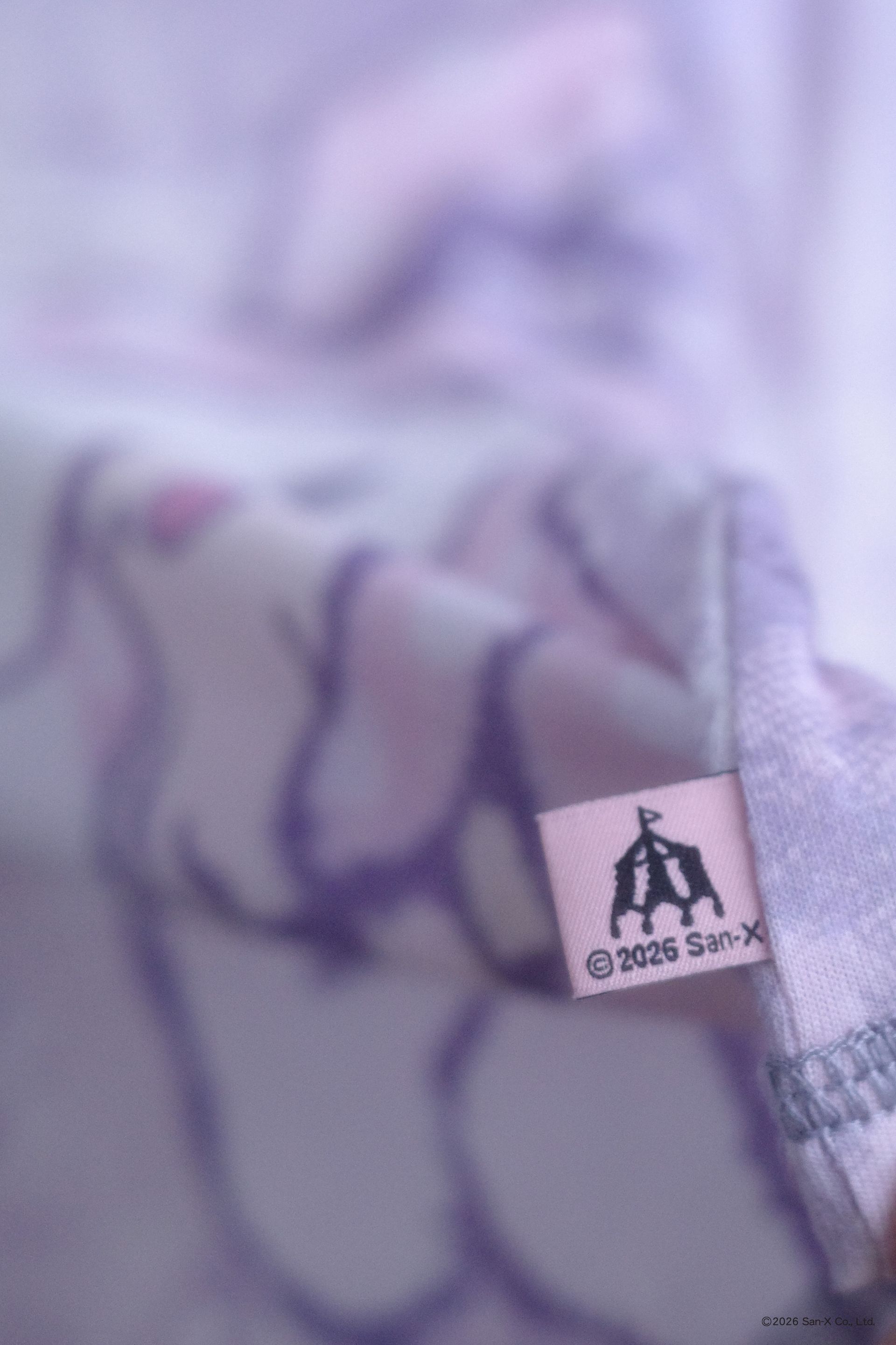 Close up image of an ethereal engineered print long-sleeve top featuring Shappo the Ringmaster. Cut from a lightweight, 110gsm 100% Cotton with chunky, heathered dusty purple ribbing at the neck and sleeve cuffs. The sleeves are finished with taller cuffs and built-in thumb holes, providing extra coverage and keeping your hands warm during the colder months. Pink tag with a circus tent printed on it.