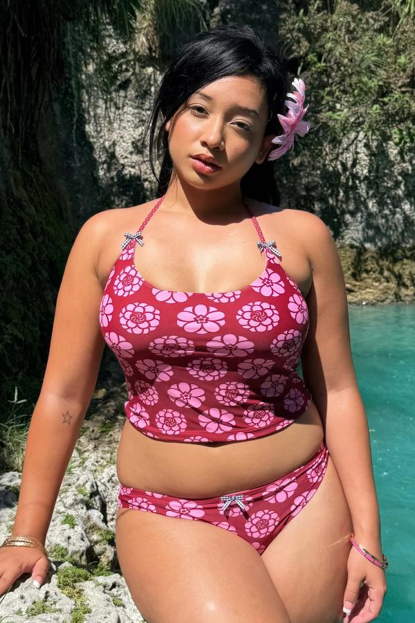 Woman wearing the Kiku Print tankini. Pattern is a maroon background with circular pink flowers. Straps have gingham bows attached. Tankini is cropped at the waist. She stands against a tropical background.