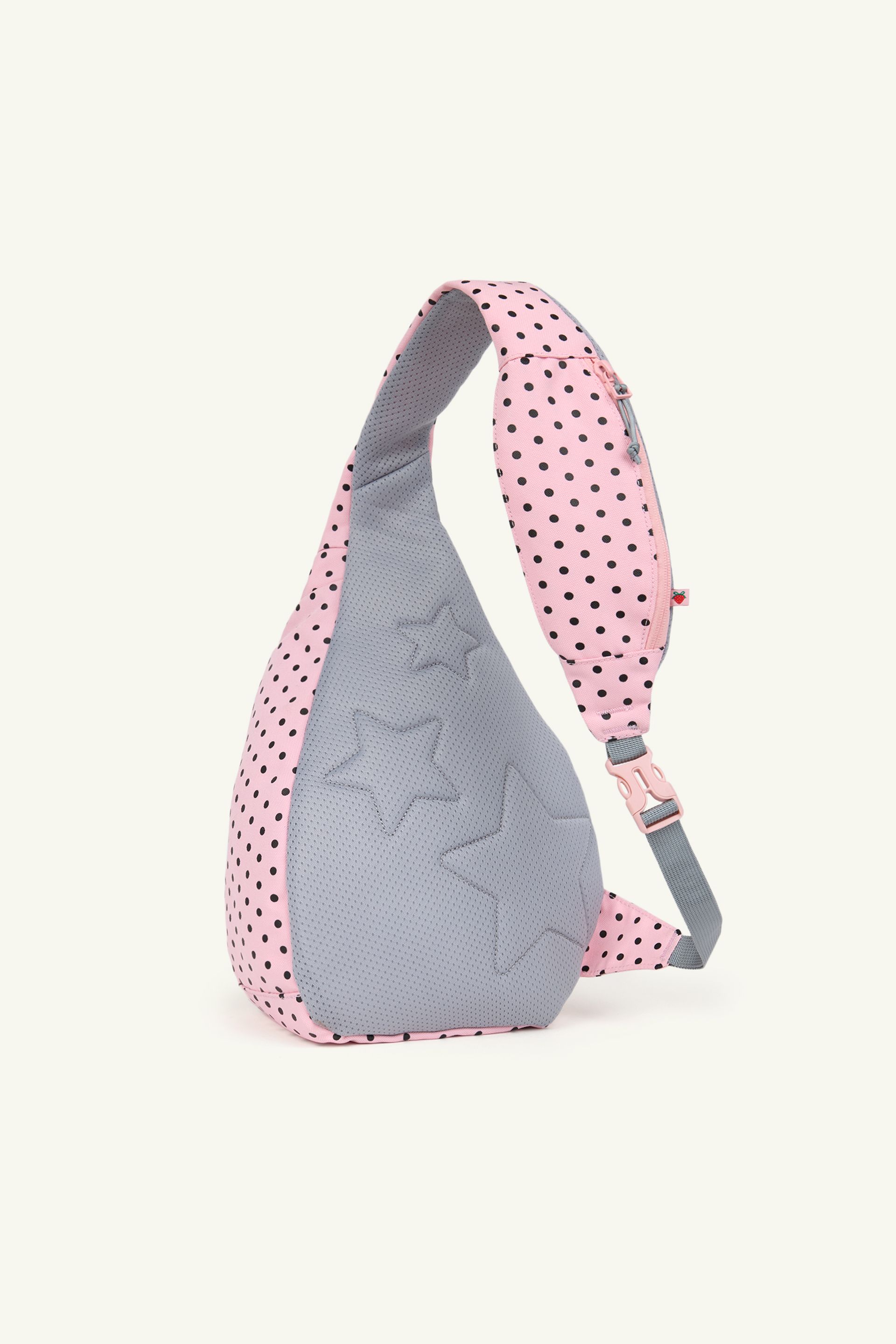 Back view of a pink sling backpack with black polka dots and a gray padded panel featuring embossed star shapes. The bag has a gray adjustable strap with a pink buckle and a small zip pocket on the strap.