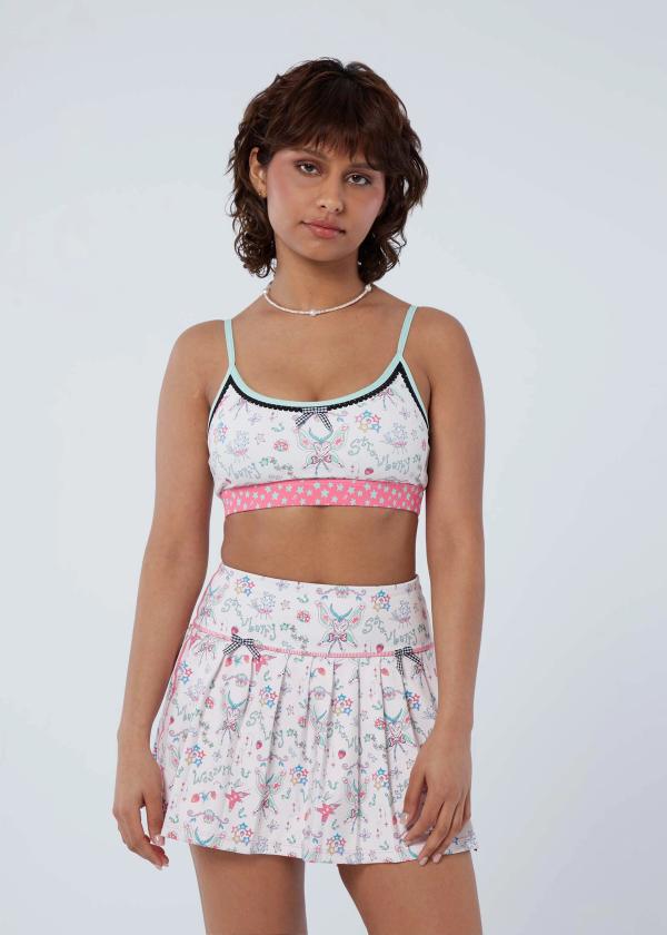 Woman with arms at sides wears a bralette silhouette sports bra in a fairy-like starbaby print with a pink and mint green shooting star waistband, black picot trimming, mint binding and a black and white gingham bow at front and a matching pleated skort.