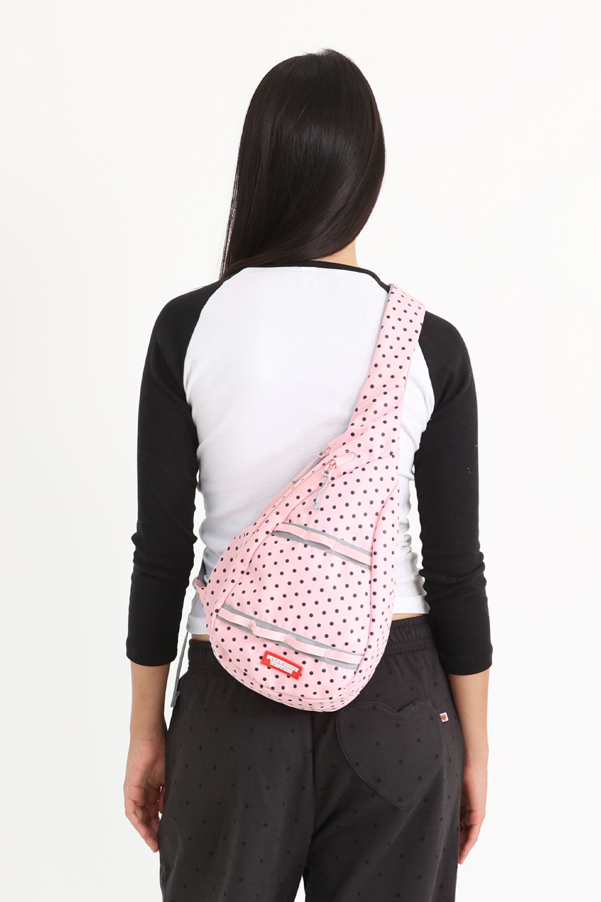 Back view of a model, She has a pink polka-dotted sling bag with gray zippers and a small red logo tag slung across her back.