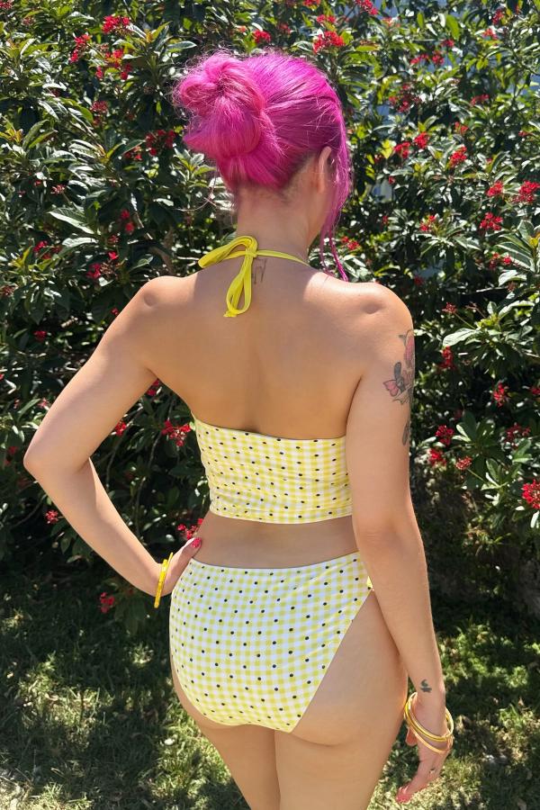 Back view of woman wearing the Yellow Gingham Print tankini. Straps have gingham bows attached. Tankini is cropped at the waist. She stands against a green bush.