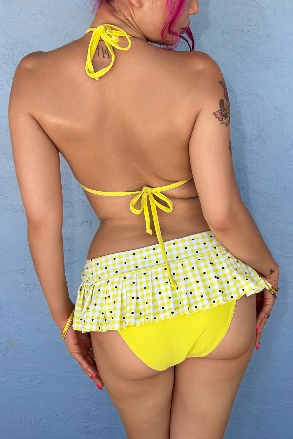 Back view of woman wearing the Yellow Gingham Print micro pleated skirt bikini bottom with attached yellow bottoms. Gingham bows at front, against a wall.