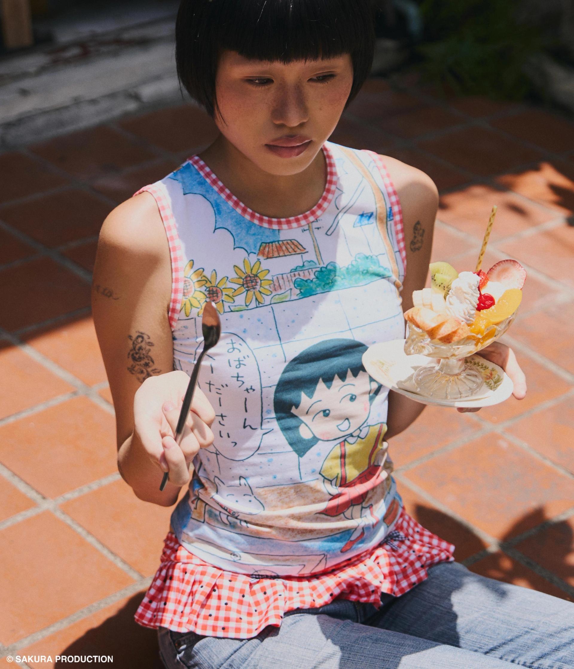 Woman sitting on stoop with a parfait wearing a tank top with a print of the original Chibi Maruko Chan manga cover art illustration.