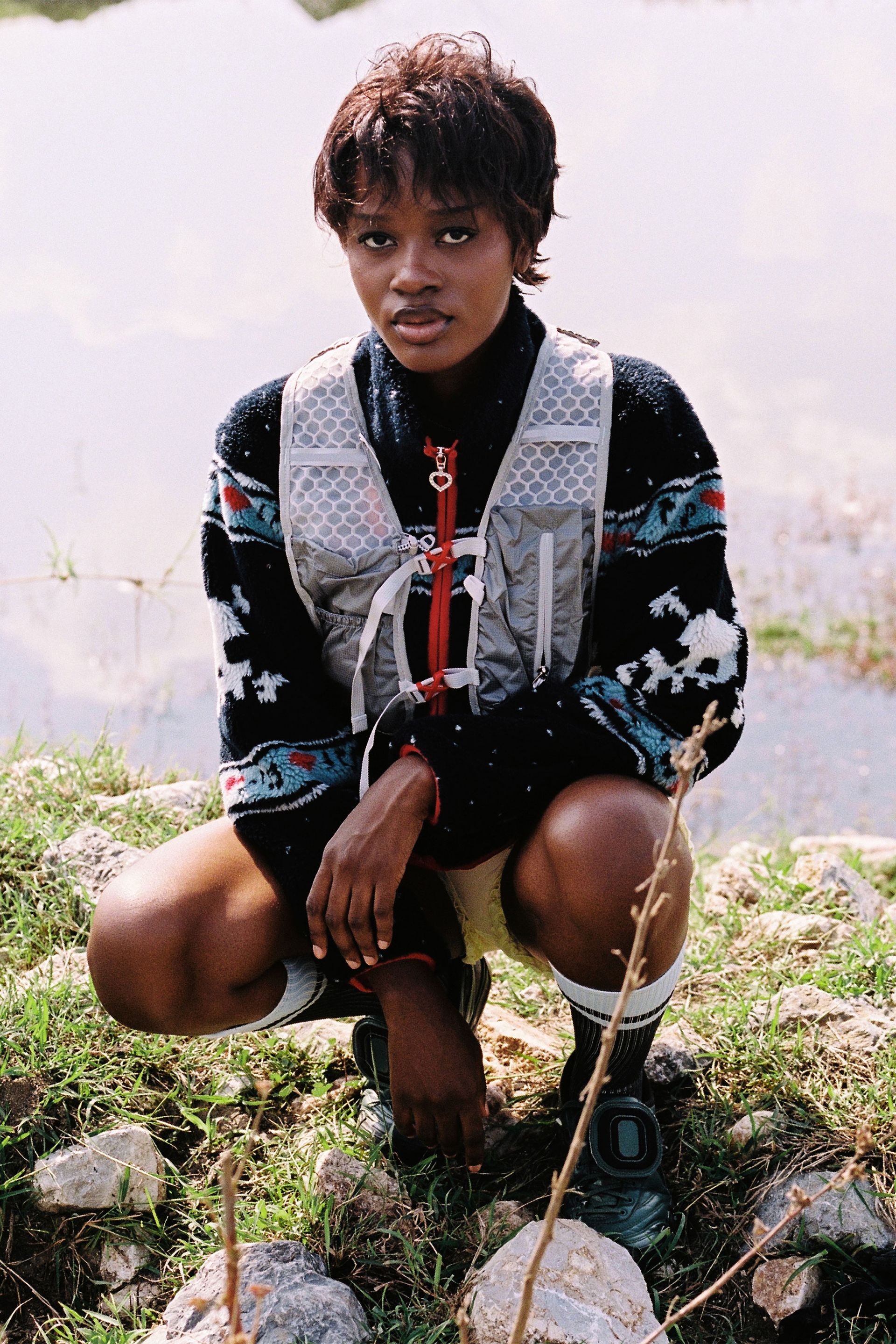 Woman squatting wears Black Multi skully sherpa jacket featuring white skull and crossbones patterns, red hearts, and light blue musical note designs across the chest and sleeves against a pond. She wears a vest over sherpa. 