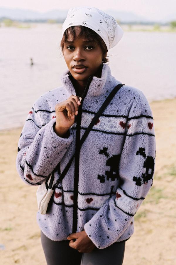 A hike to a lake, zipping up the Dusty Purple sherpa jacket featuring skull and crossbones patterns, dark red hearts, and light purple musical note designs across the chest and sleeves. 