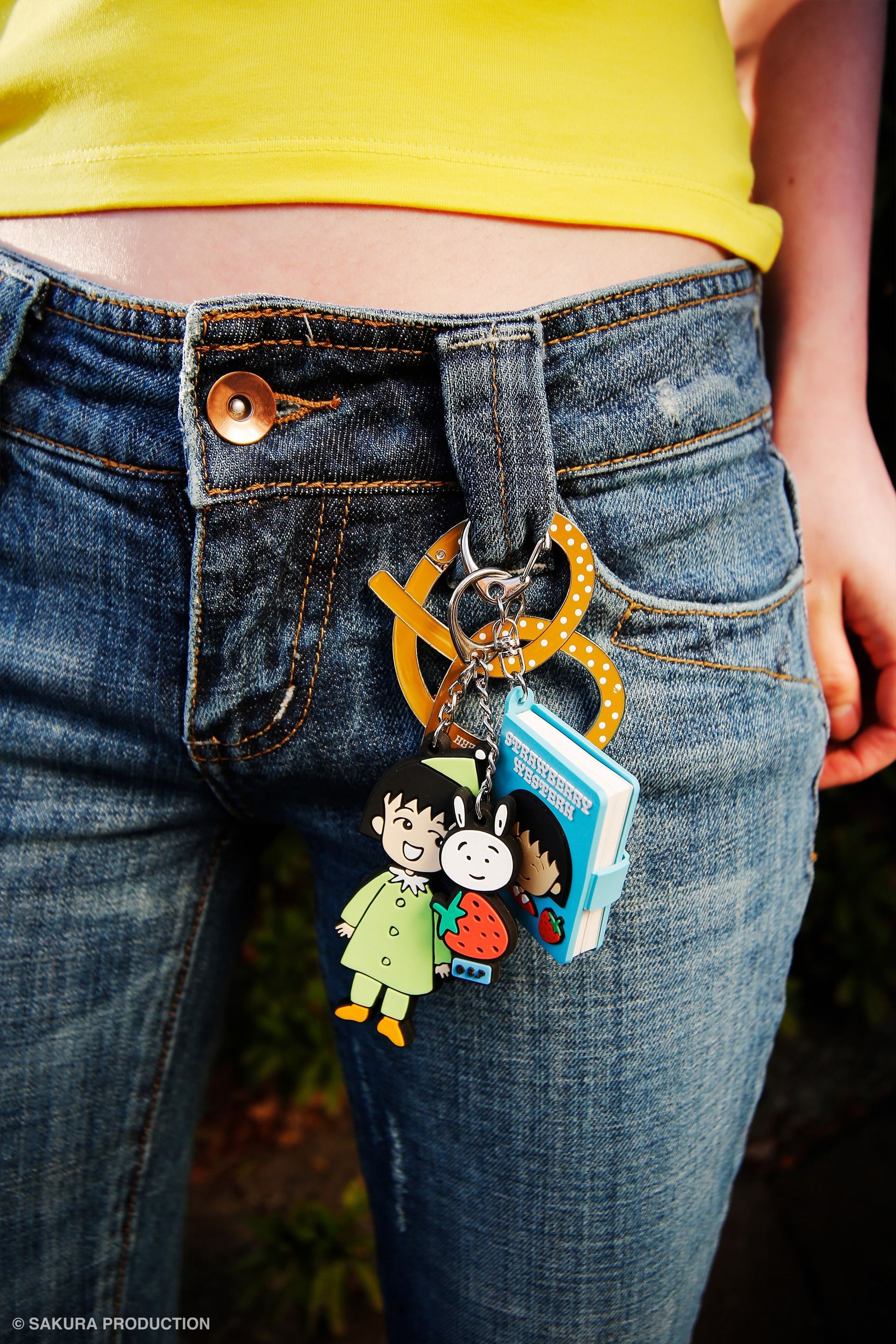 Chibi Maruko Chan Tomodachi Keychain hanging from the belt loop of someone's jeans. Features Chibi Maruko-chan wearing a festive, green outfit on one chain. On the other chain is her bunny friend stacked on top of a strawberry. 
