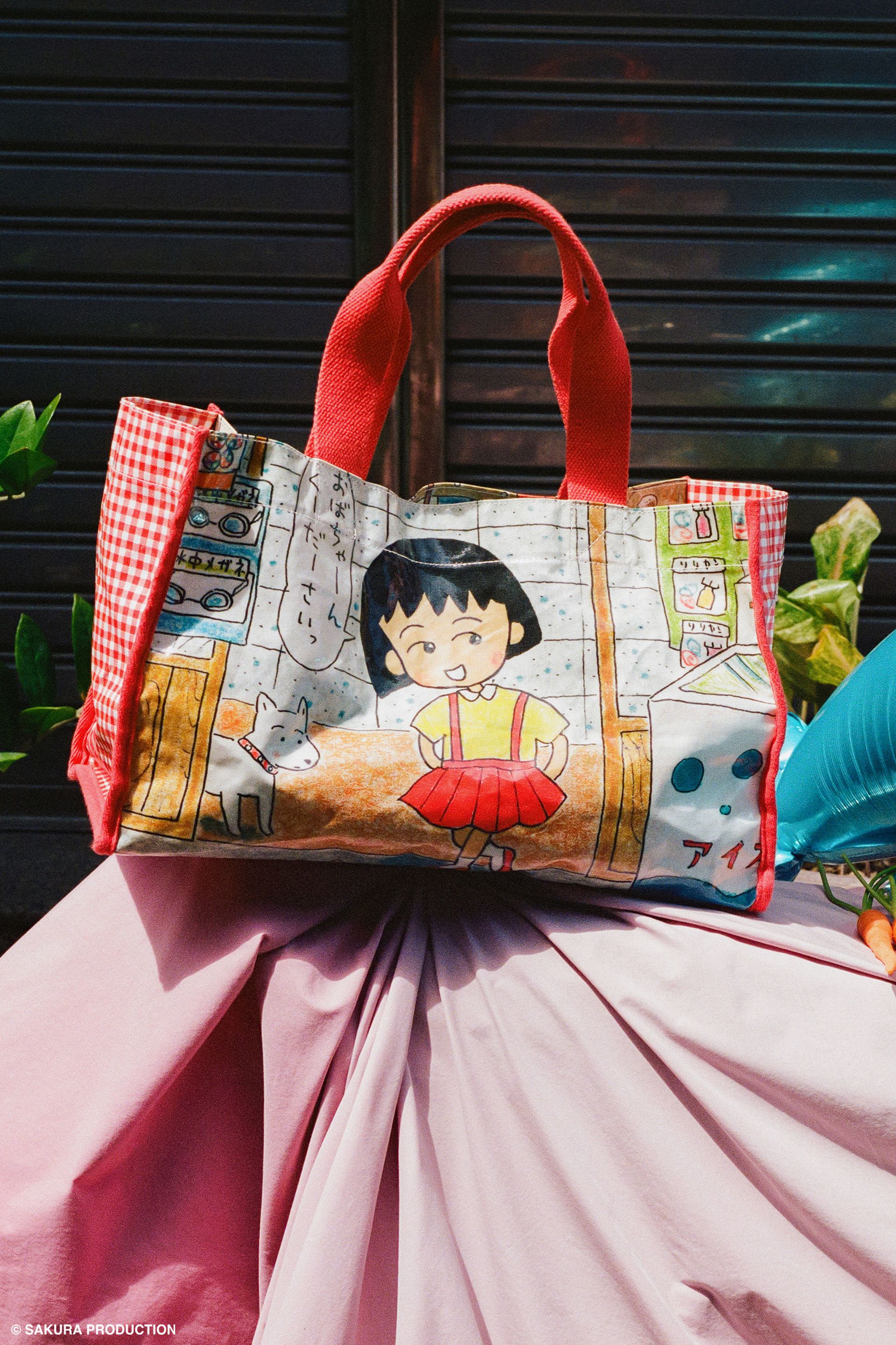 Carryall Bag sitting on top of pink cloth against a shuttered store. Shows image of manga drawing Chibi Maruko Chan and her dog walking into a store. She says "Grandma Please" in Japanese. Side panels red gingham print. Handles and trim are red. 