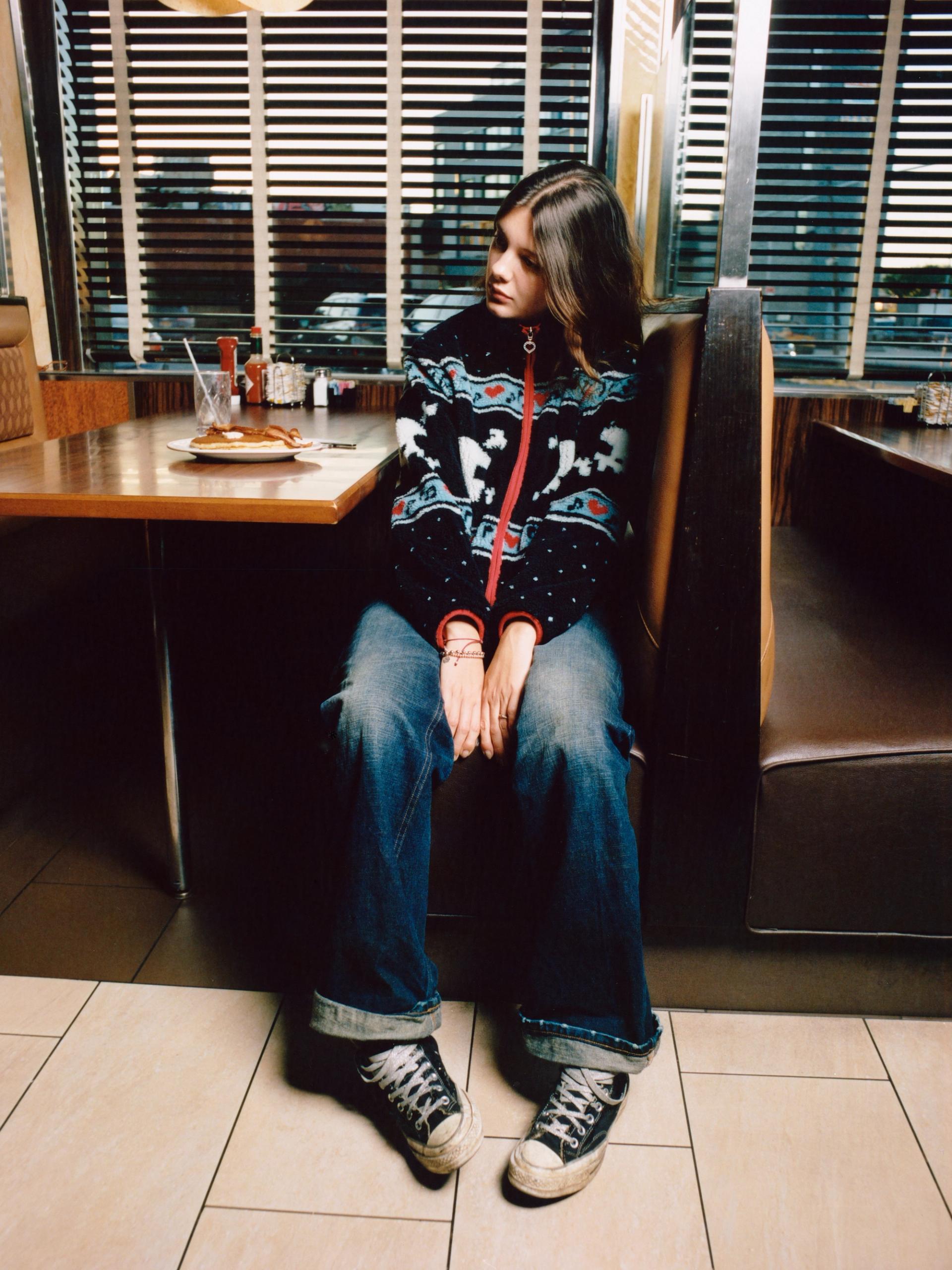 Model sits in a diner booth wearing a black multi sherpa jacket with red trim and a pattern of white skulls, hearts, and blue geometric designs.