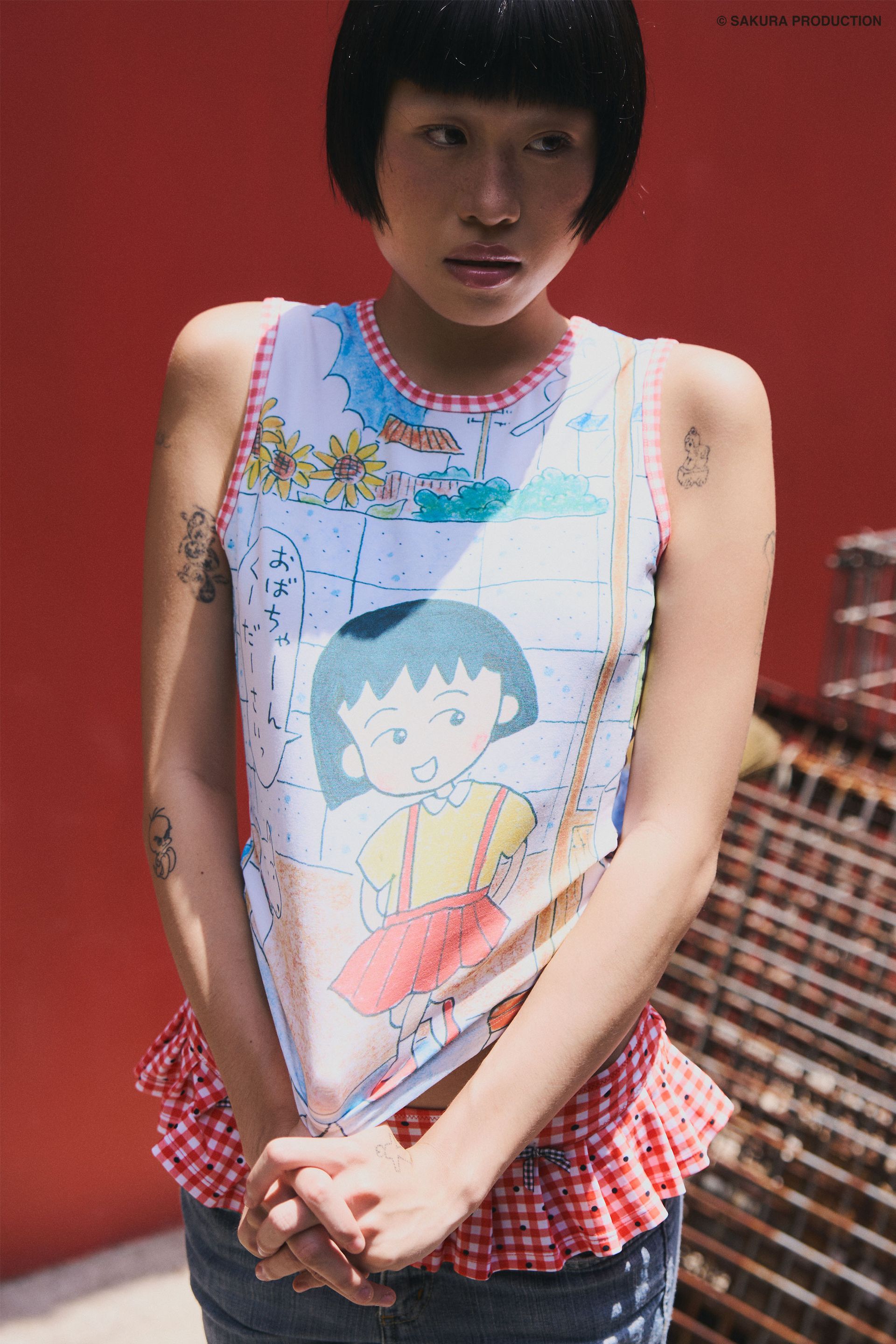Woman wearing and tugging at Sweet Shopper Tank. Top illustrates a manga-style image of a young girl and her dog walking into a store. She speaks in Japanese, "Please Grandma!" The top has a red gingham trim around the neck and sleeves. She wears jeans and a red gingham swim bottom over them against a red background with crates.