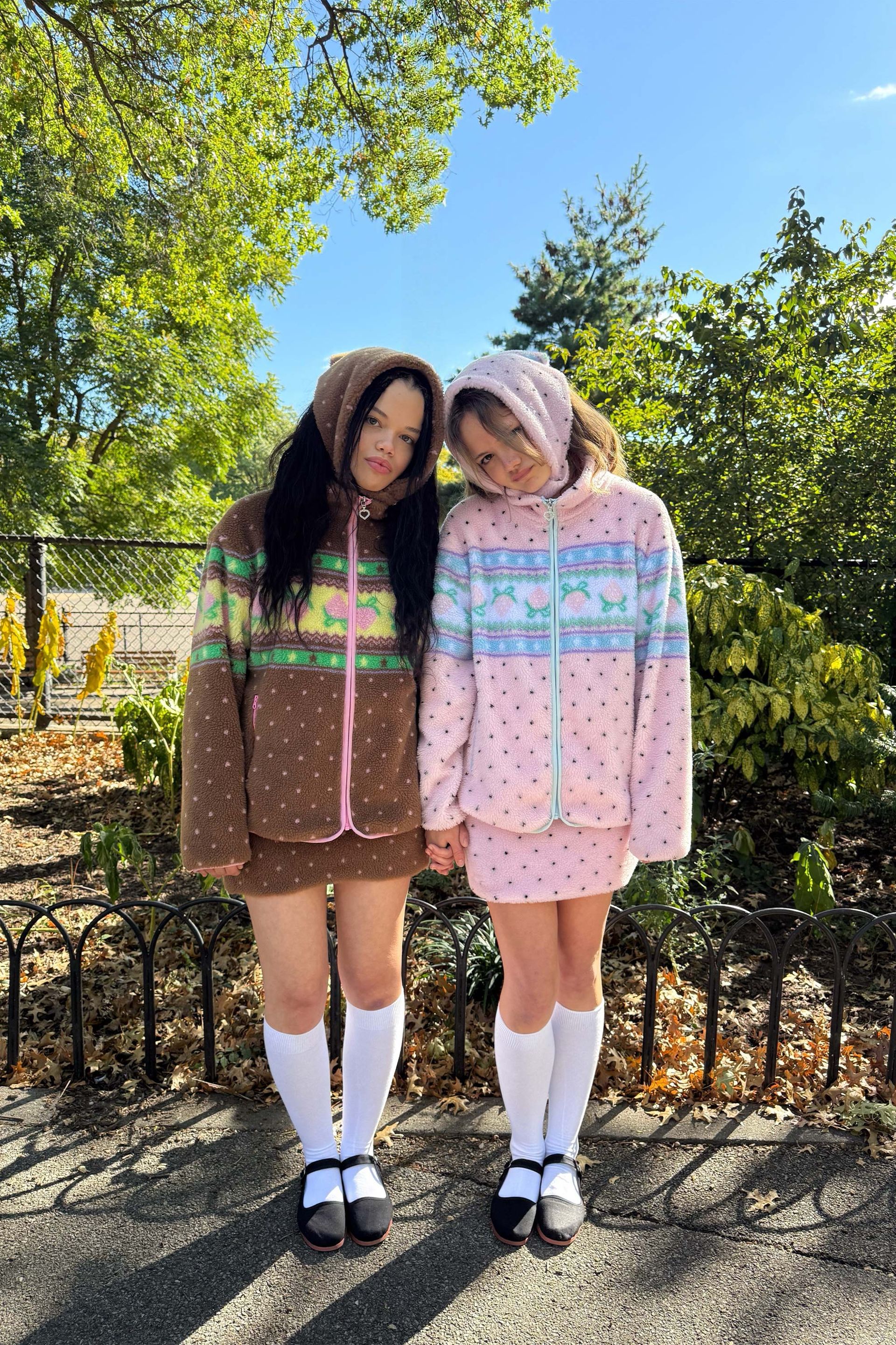 Each model wearing our cozy choco sherpa jacket with a dotted pattern and pastel design. One wears our Brown choco jacket with green and pink accents, while the other wears the Pink version with blue detailing.