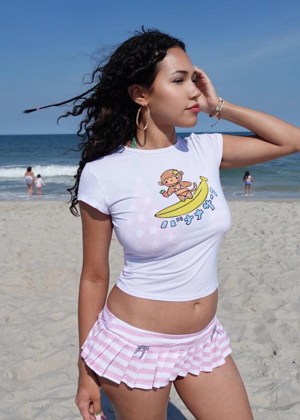 Girl standing on sand at beach wearing a baby tee with a surfing monkey graphic and a pink and white border stripe micro skirt bikini bottom.