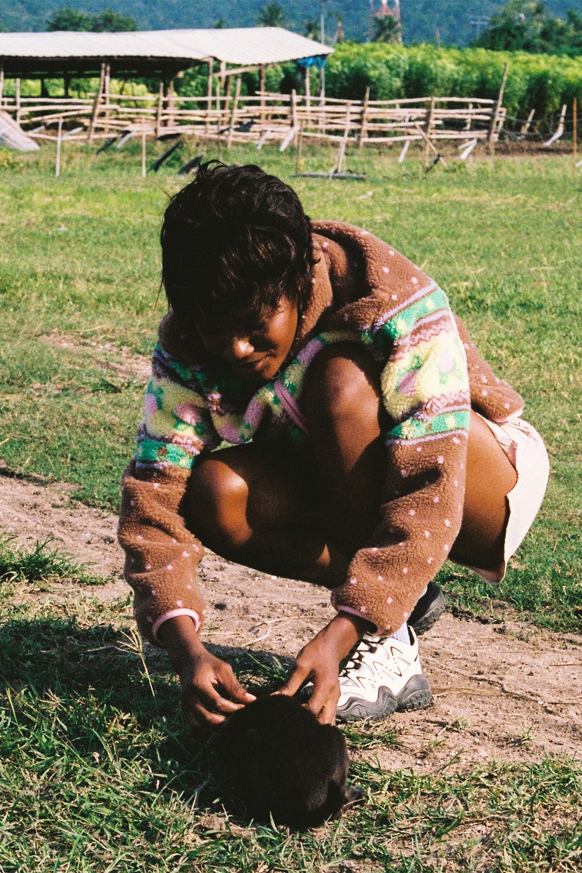 Woman petting a puppy at a farm in the Brown fluffy jacquard knit sherpa jacket in a choco strawberry border motif with binding, zippers and a metal heart shape zipper pull.