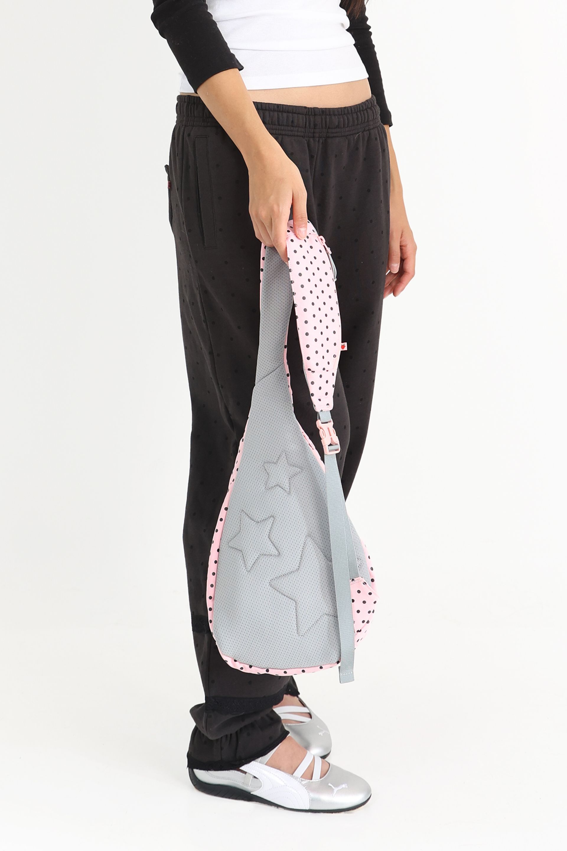 Model holds a pink polka-dotted sling crossbody bag by the strap at their side, showing the gray mesh back panel with embossed star shapes.