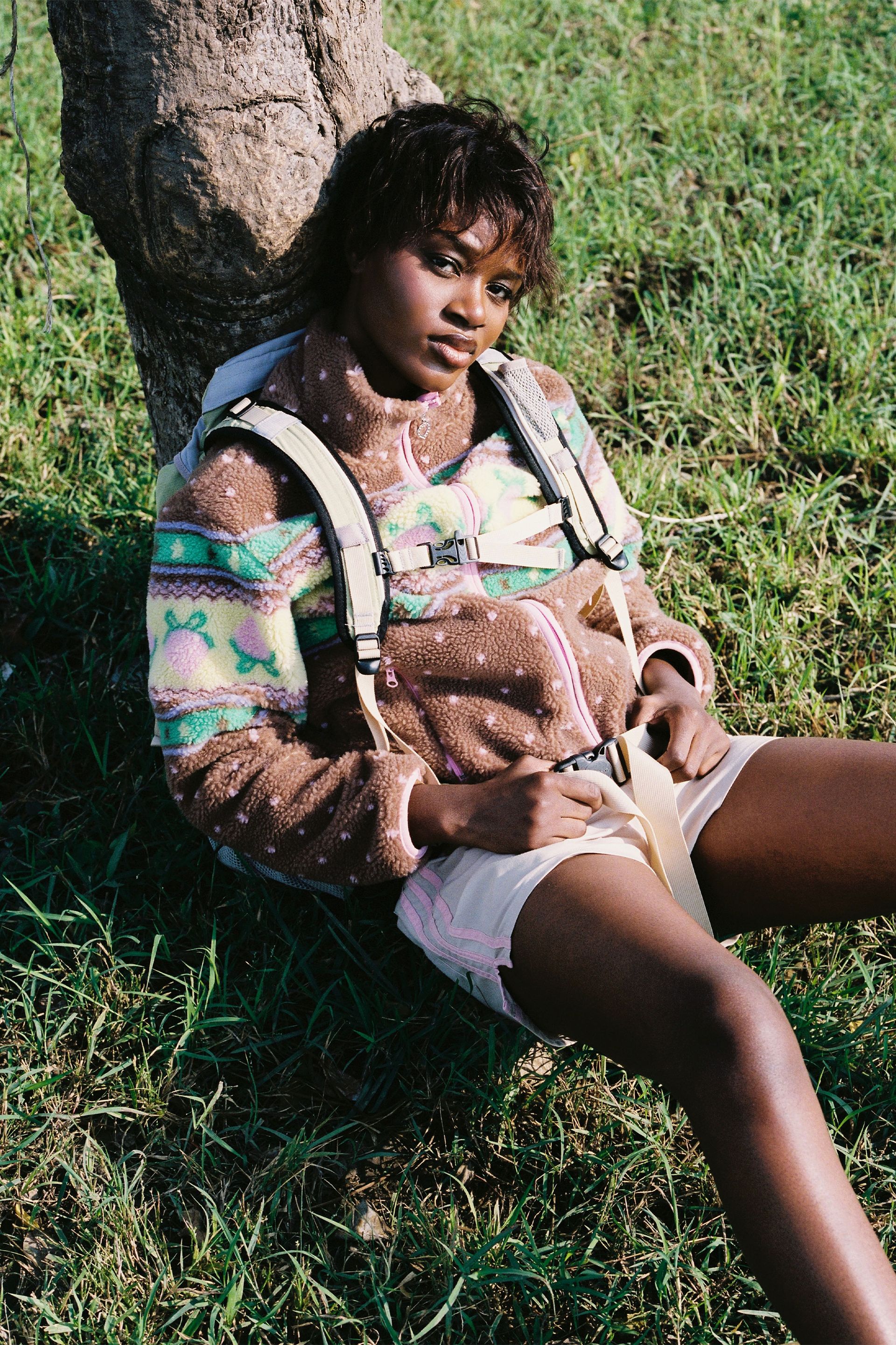 Sitting down leaning against a tree trunk in the Brown fluffy jacquard knit sherpa jacket in a choco strawberry border motif with binding, zippers and a metal heart shape zipper pull.