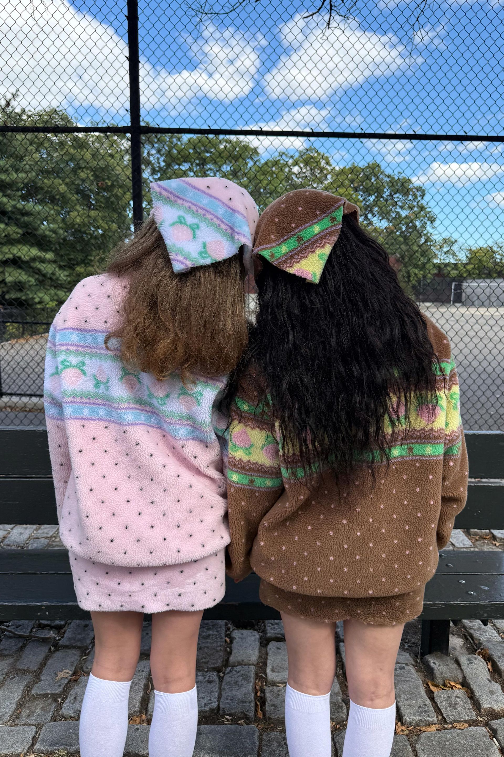Back view of two models wearing the pink sherpa jacket with black polka dots and pastel blue and green strawberry-inspired stripes across the back. Second model is wearing the brown sherpa jacket with pink polka dots.