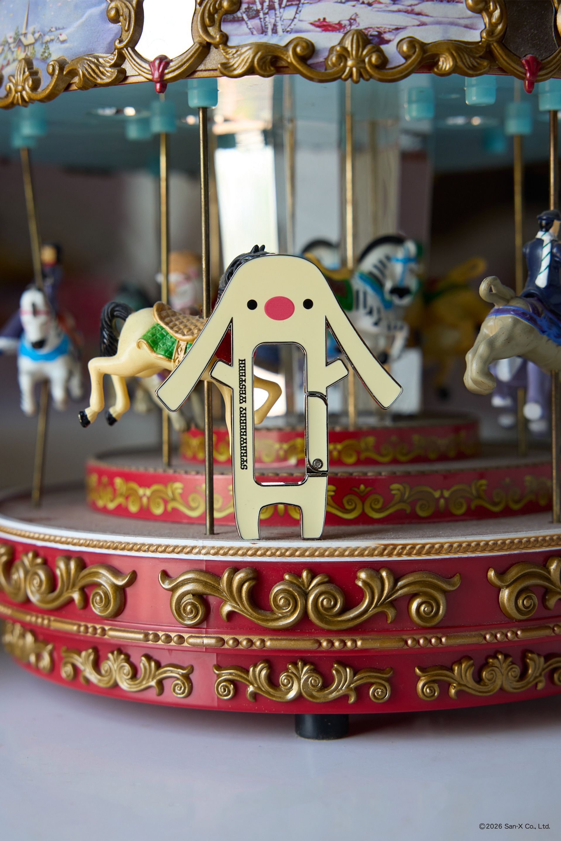 Toto carabiner standing on a mini carousel. He is shaped like a dog/bunny hybrid with long, floppy ears and a big red nose. The skin color is yellow with two black dots for eyes. Strawberry Western logo on the front.