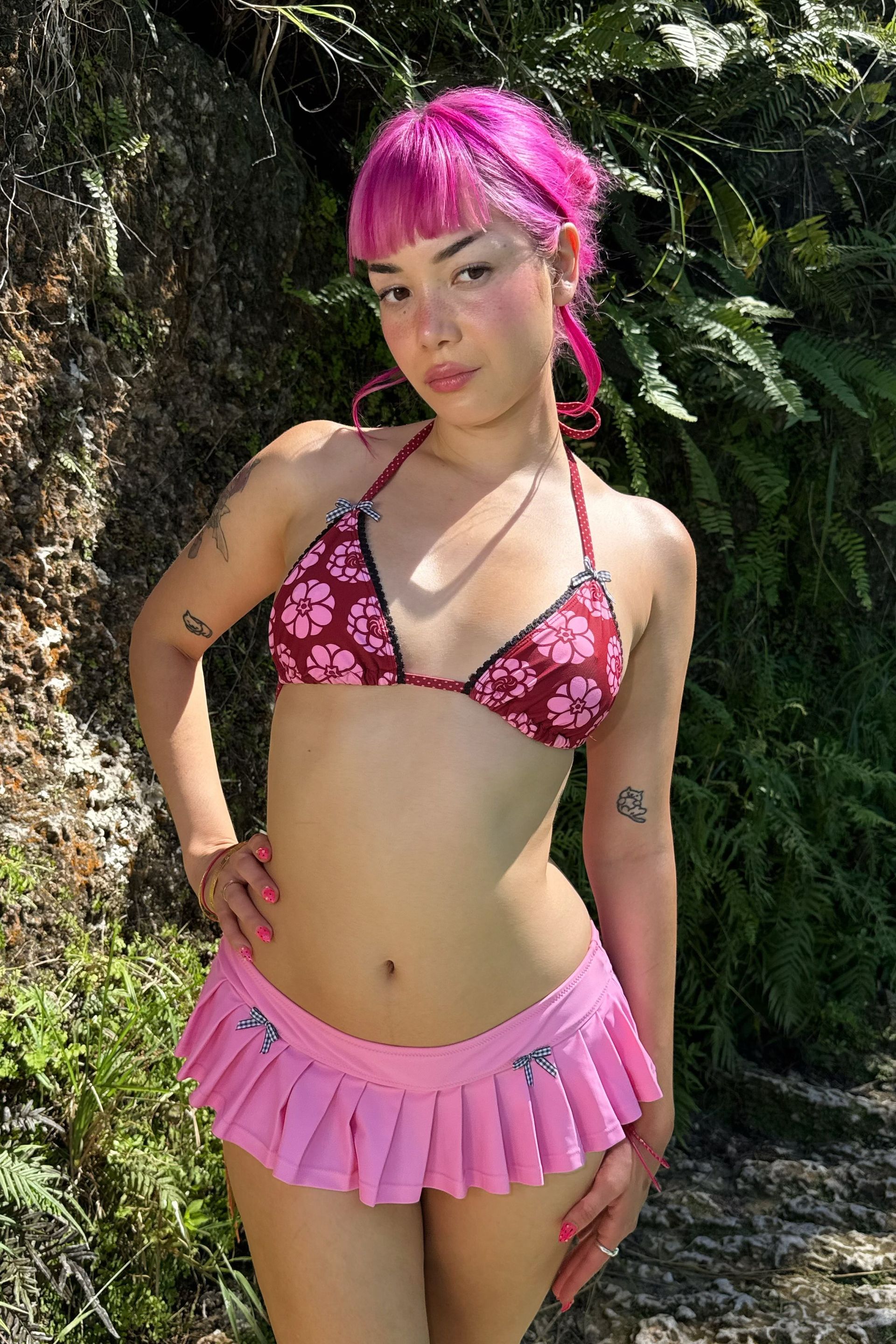 Woman wearing the Kiku Print triangle bikini top. Pattern is a maroon background with circular pink flowers. Each cup is adorned with black scallop lace trim and gingham bows. She stands against a natural background.