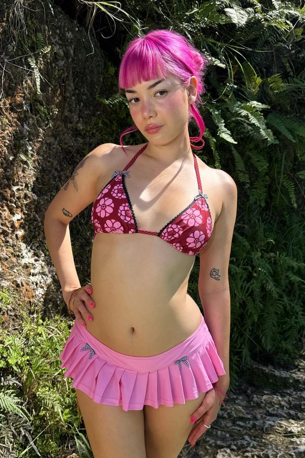 Woman wearing the Kiku Print triangle bikini top. Pattern is a maroon background with circular pink flowers. Each cup is adorned with black scallop lace trim and gingham bows. She stands against a natural background.