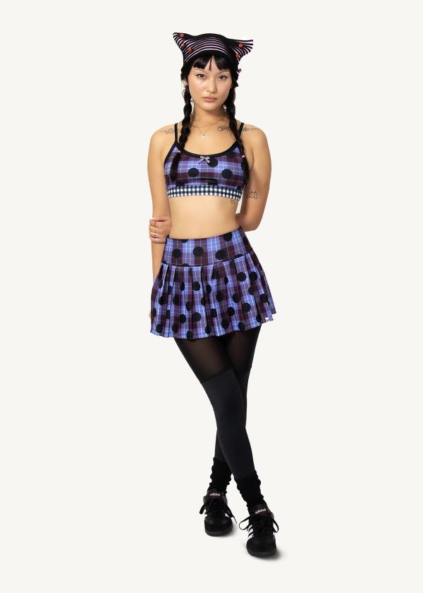 Model stands with one arm behind back wearing a sports bra and a stretchy pleated school girl skirt in a plaid polka dot print with attached interior legging.