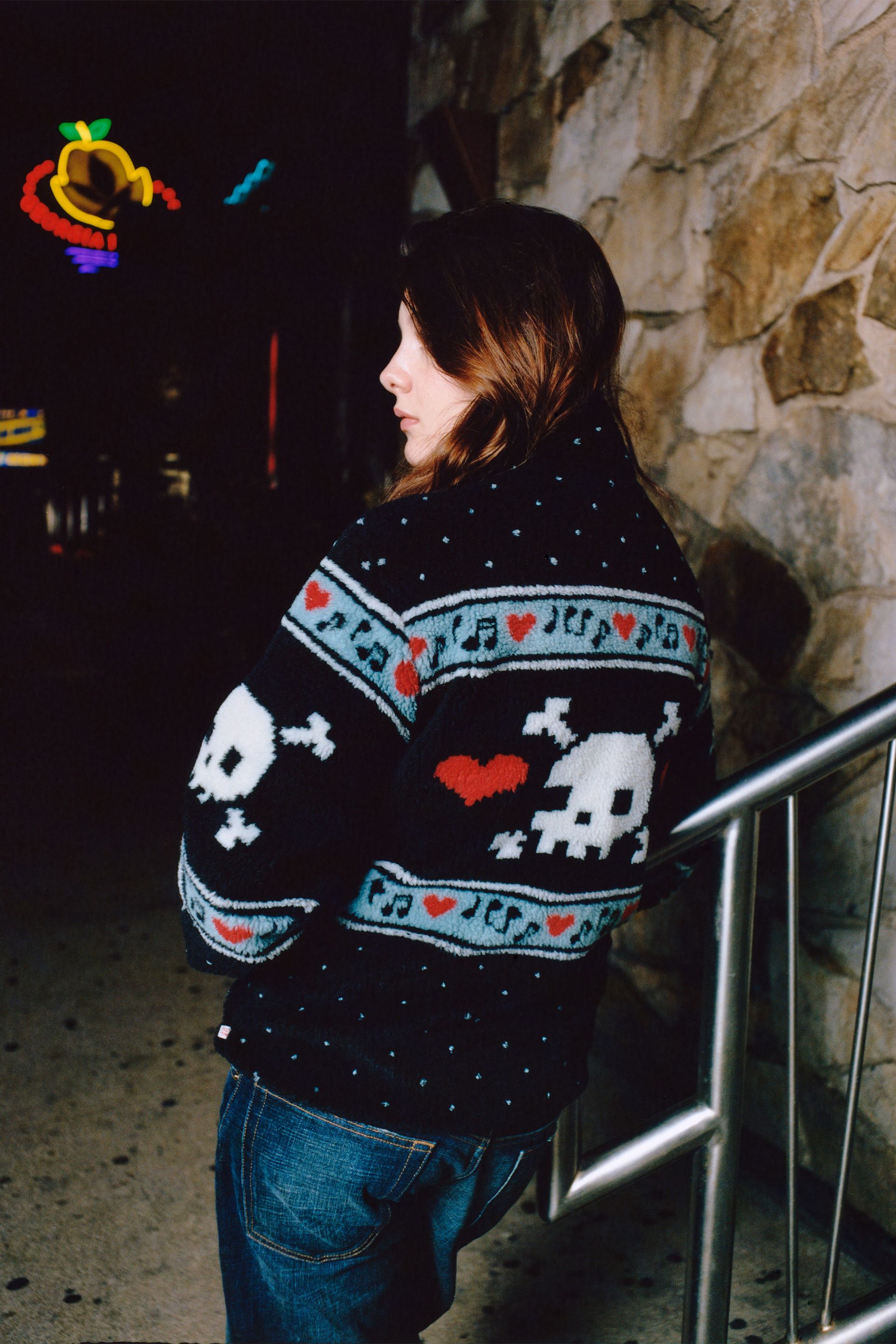 Model is seen from the side and back. She wears a black multi sherpa jacket featuring white skull and crossbones motifs, red hearts, and blue decorative patterns.