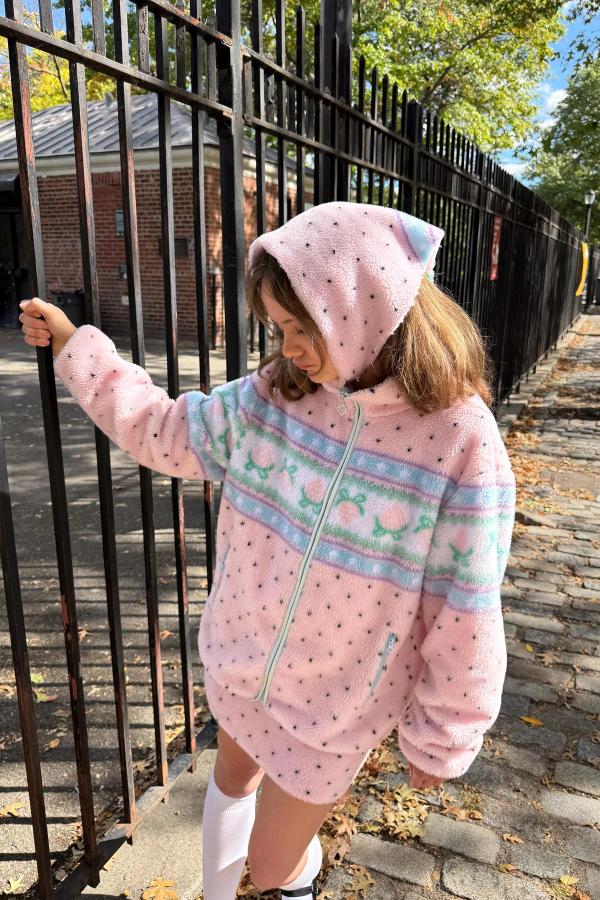 Model stands outdoors wearing a Pink sherpa jacket with black polka dots and a baby blue stripe pattern across the chest. The jacket features a front zipper and pockets.