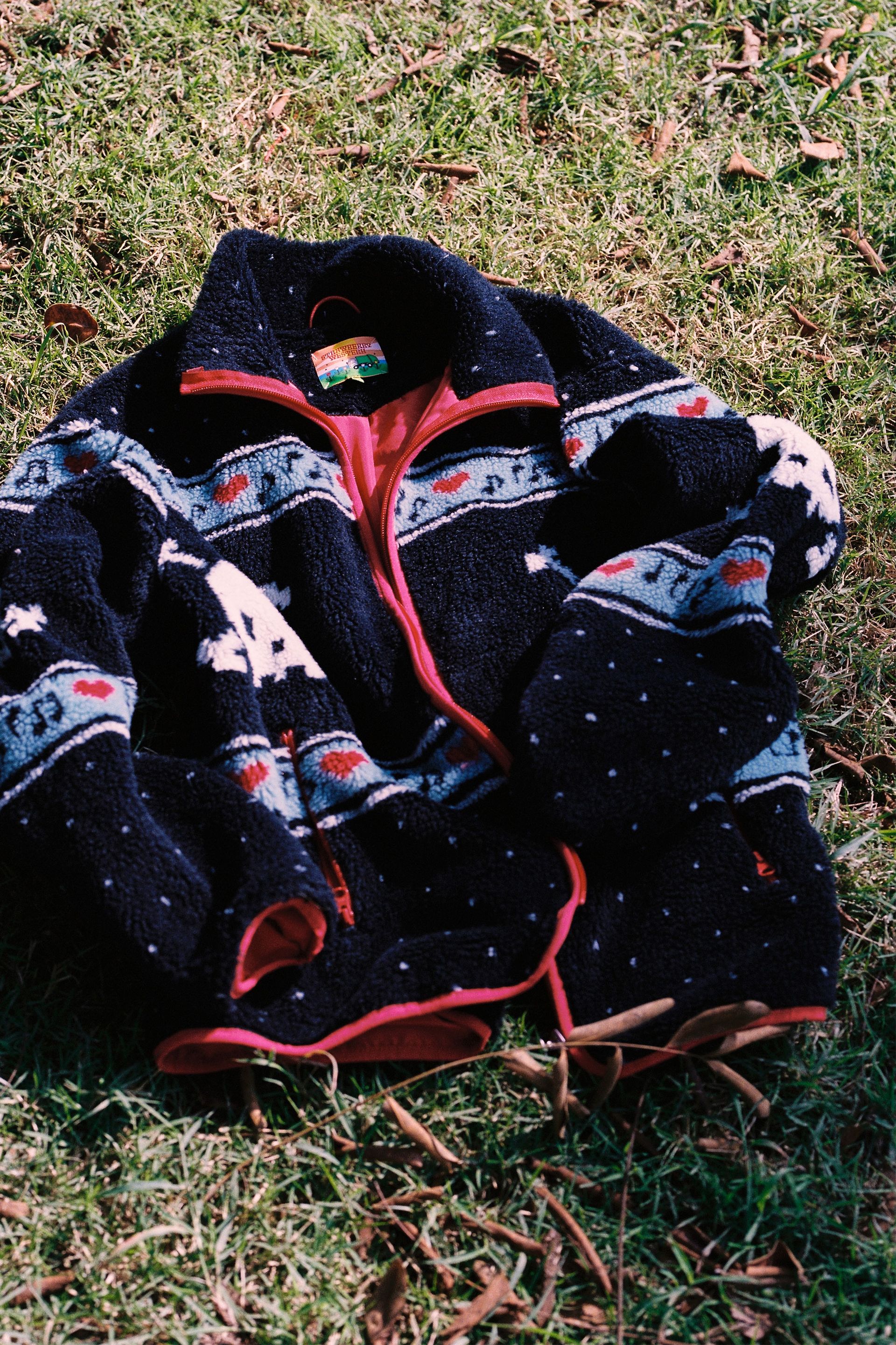 Black Multi skully sherpa jacket featuring white skull and crossbones patterns, red hearts, and light blue musical note designs across the chest and sleeves laying in grass
