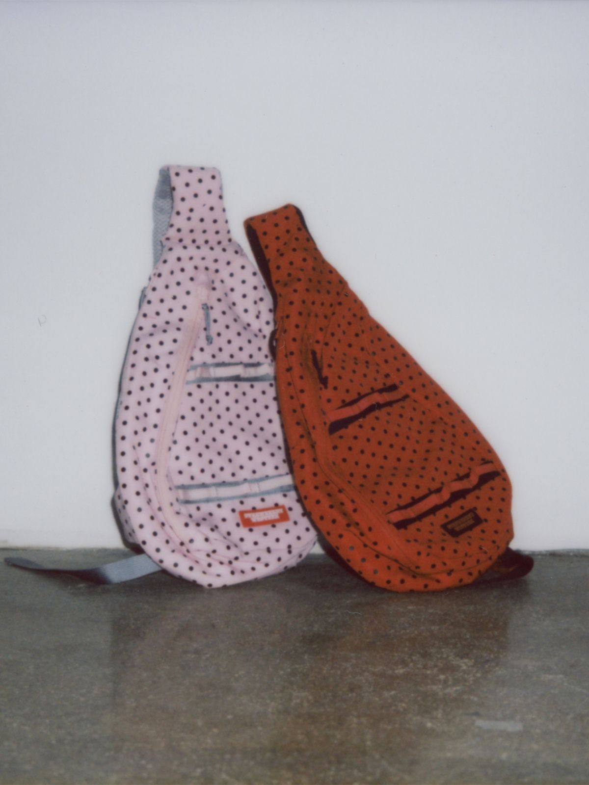 Polaroid photo of a pink Sling Bag and Red Sling Bag on the floor, leaning up against a white wall.