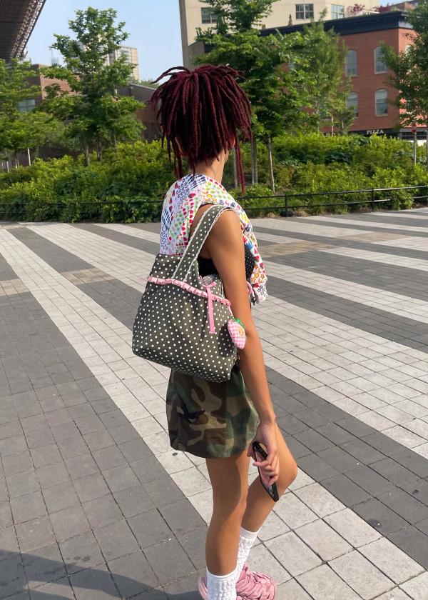 Girl walking on the street wearing an olive color polka dot ruffle trim shoulder bag with crochet scallop edging, a pink bow and a strawbaby keychain attached.