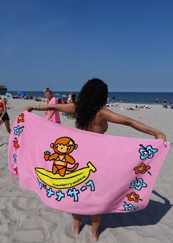 Girl at beach holding up kawaii 100% Cotton, 400gsm beach towel with digitally printed surfing monkey artwork with 'Banana Surf' written in Japanese.