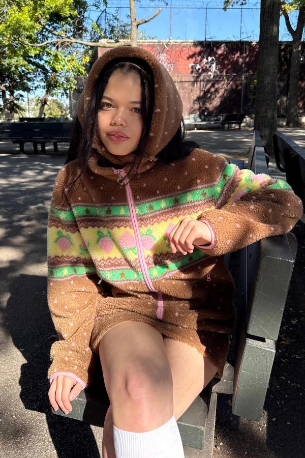 Model sits outdoors on a park bench wearing a cozy Brown sherpa jacket with a pink zipper and pastel green and yellow stripe pattern featuring strawberries.