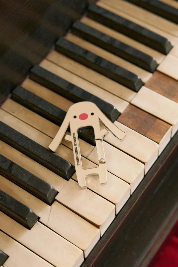 Toto carabiner laying on piano keys. He is shaped like a dog/bunny hybrid with long, floppy ears and a big red nose. The skin color is yellow with two black dots for eyes. Strawberry Western logo on the front.