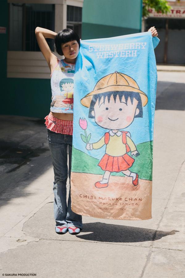 Woman standing holding towel in the street. Towel shows hand drawn image of young Chibi Maruko Chan holding a pink tulip against a natural backdrop. She wears the Sweet Shopper Tank, jeans, and a red gingham skirt bikini bottom.