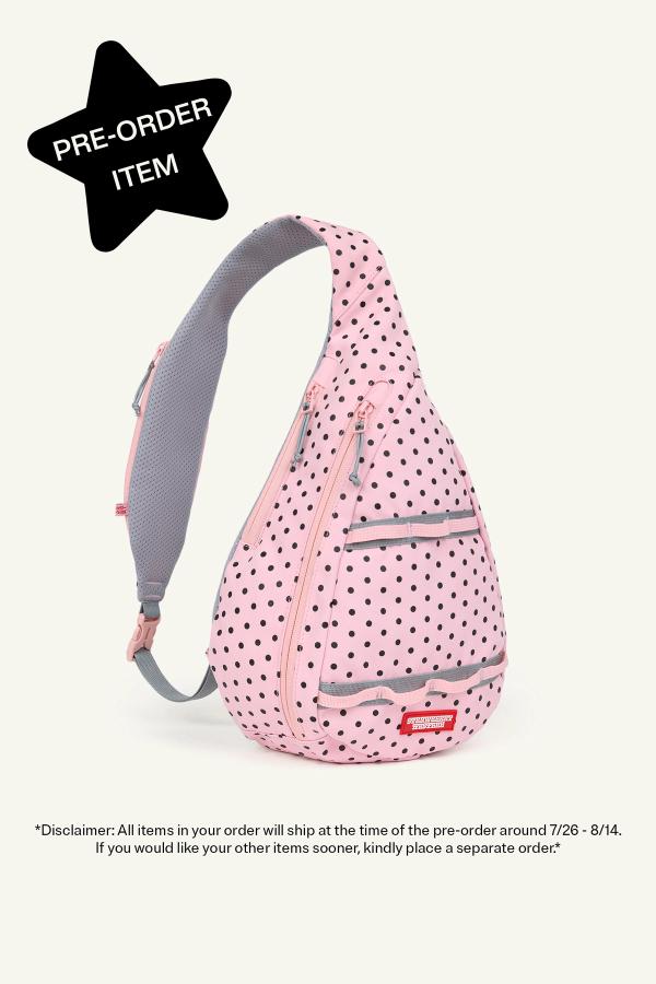 Product image of front view of a Pink sling backpack with black polka dots, gray zippers, and a gray adjustable strap. The bag has multiple front zip compartments. In the top left corner a black star reads "Pre-Order Item." Bottom reads “Disclaimer: All items in your order will ship at the time of the pre-order around 7/26-8/14. If you would like your other items sooner, kindly place a separate order.