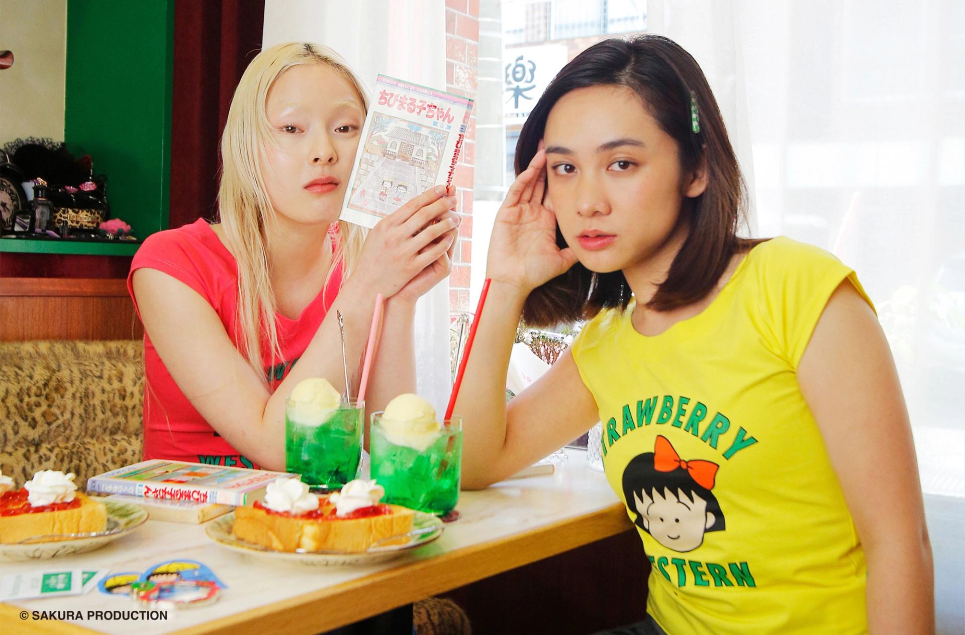 Two girls sitting in a dinner wearing the Chibi Maruko Chan Raglan tops. The girl on the left is reading manga.