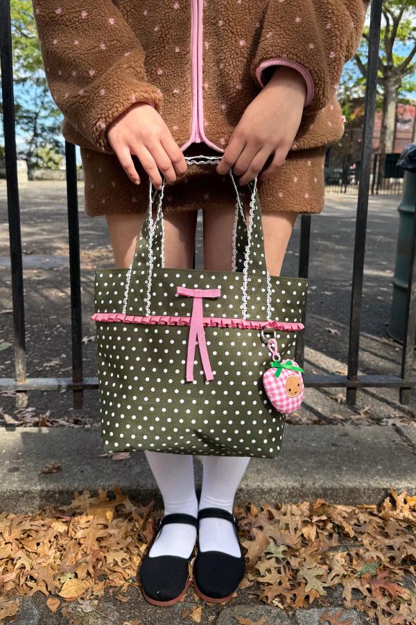 Person standing outdoors holds a small olive-green tote bag with white polka dots, trimmed with a pink ribbon and lace detail.