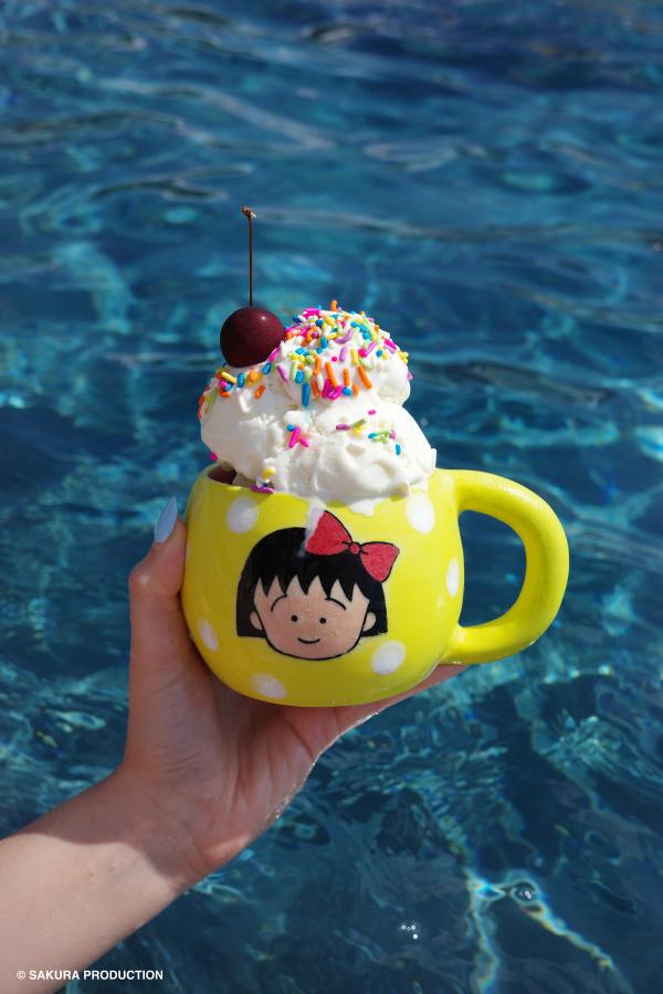 Chibi Maruko Chan hand made ceramic mug held against a pool backdrop. Yellow body and white polka dots with face of a little girl with a red bow in her hair. There is ice cream with rainbow sprinkles and a cherry in the mug.