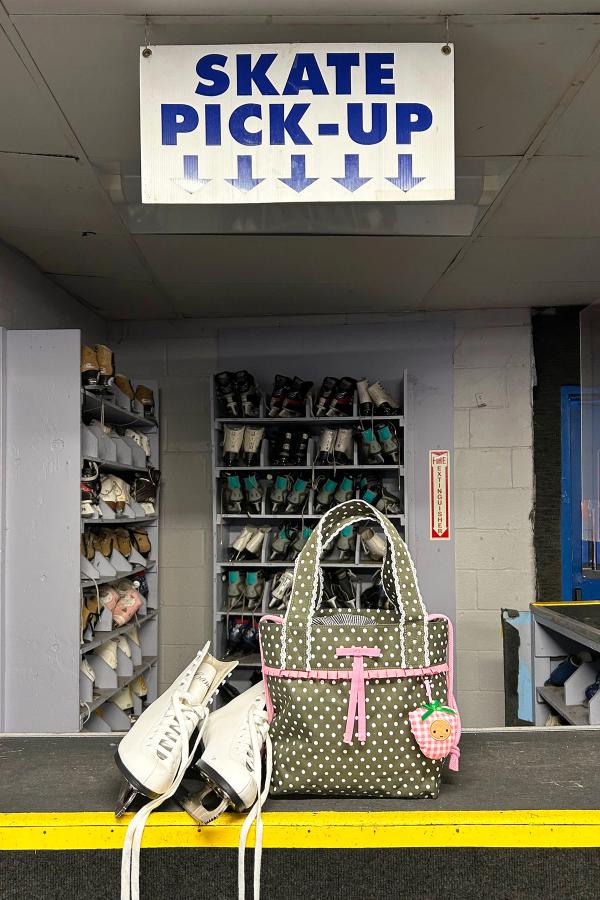 An olive color polka dot ruffle trim mini shoulder bag with crochet scallop edging, a black and white gingham lining and a drawstring cinch closure sits on the counter of a skate rental..