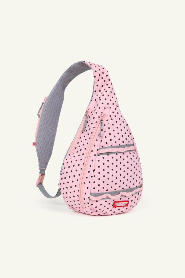 Front view of a pink sling backpack with black polka dots, gray zippers, and a gray adjustable strap. The bag has multiple front zip compartments.