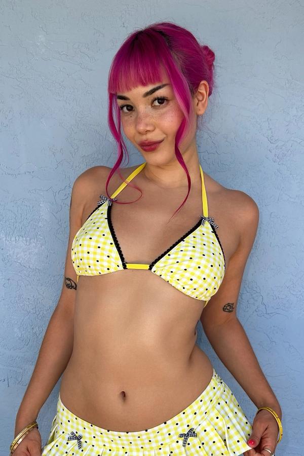 Woman wearing the Yellow Gingham Print triangle bikini top. Each cup is adorned with black scallop lace trim and gingham bows. She stands against a wall.