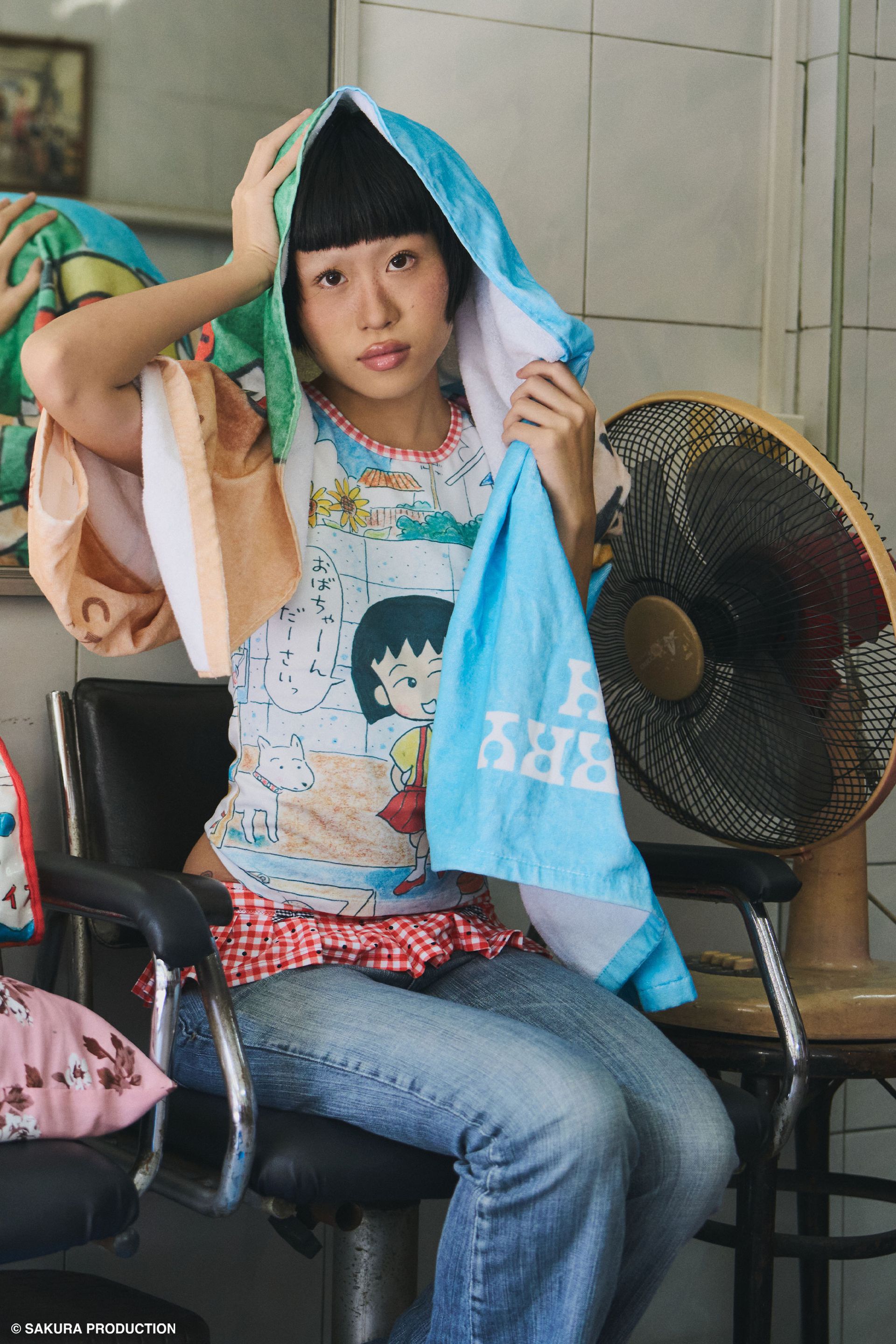 Woman sitting in a hair salon with towel draped over her head. Towel shows hand drawn image of young Chibi Maruko Chan holding a pink tulip against a natural backdrop. She wears the Sweet Shopper Tank, jeans, and a red gingham skirt bikini bottom.