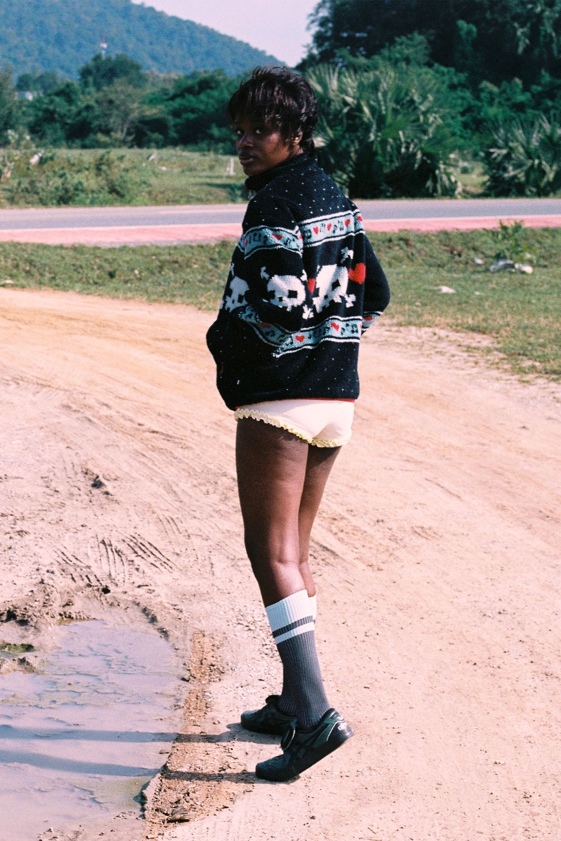 Woman from behind wears Black Multi skully sherpa jacket featuring white skull and crossbones patterns, red hearts, and light blue musical note designs across the chest and sleeves against a natural backdrop. 