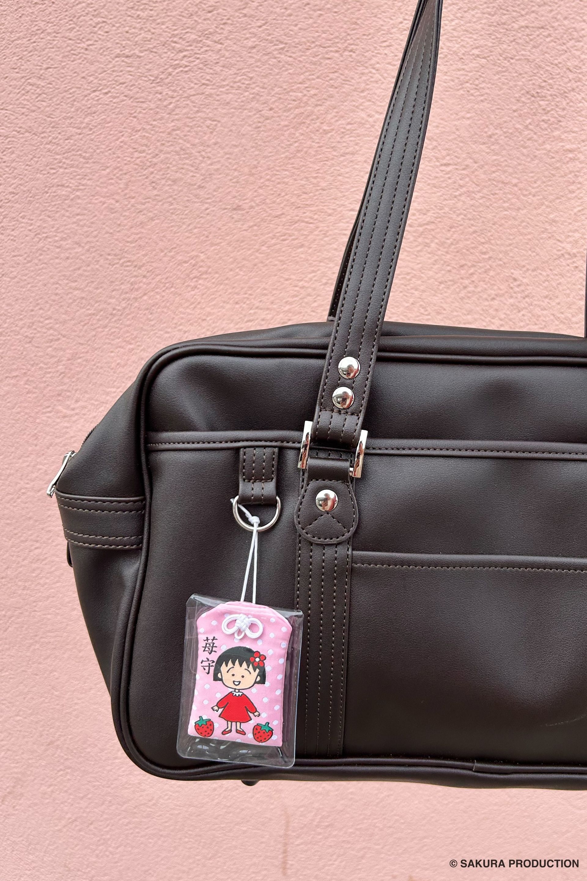 Chibi Maruko Chan omamori hanging off a messenger bag posed against a pink wall.