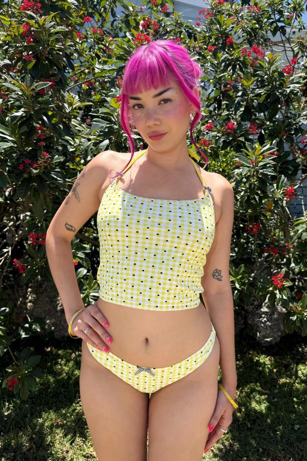 Woman wearing the Yellow Gingham Print tankini. Straps have gingham bows attached. Tankini is cropped at the waist. She stands against a green bush.