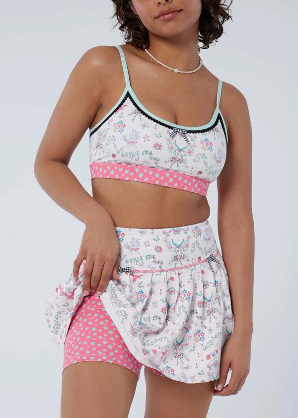 Detail view of woman pulling up pleated workout skort in a fairy-like starbaby print with gingham bows at waist to reveal the pink and mint green shooting star print attached interior shorts.