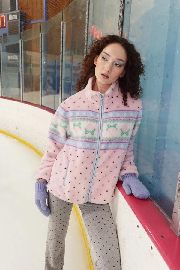 Model stands on ice rink wearing baby pink fluffy jacquard knit sherpa jacket in a strawberry border motif with blue binding.